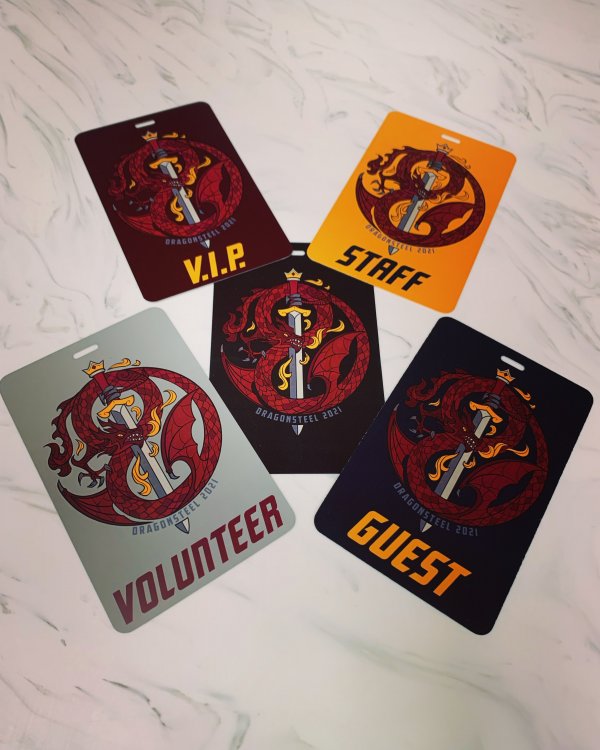Five badges sit on a table, one in each corner and one in the center. Each features a dark red dragon wrapped around a sword, with small grey text saying "Dragonsteel 2021" beneath it. In addition, all but the center badge each have a label in all caps. The top left is a VIP badge, on a darker red background and with a yellow label. The top right is a staff badge with a golden background and black label. Bottom left is a volunteer badge with a silver background and dark red label. Bottom right is a guest badge with a black background and yellow label. The center badge has a black background but no label.